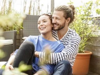 Frau lehnt sich an ihren Mann auf dem Balkon und schaut mit ihm lächelnd in die Ferne